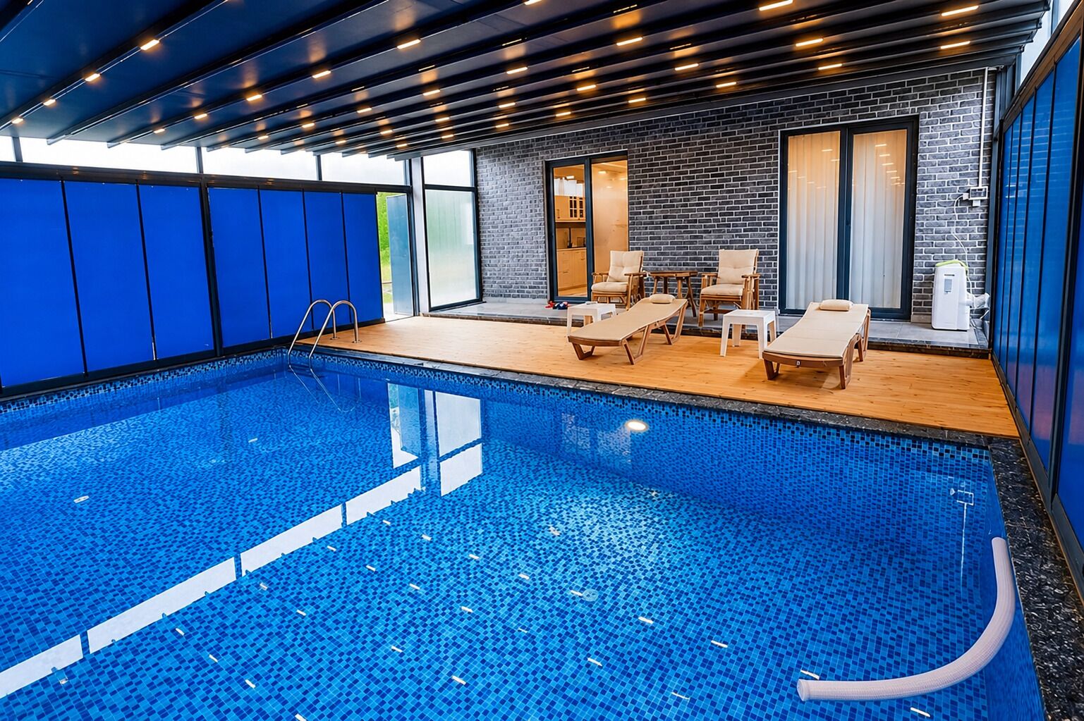 An indoor swimming pool with clear blue water, wooden deck, lounge chairs, and a brick wall.