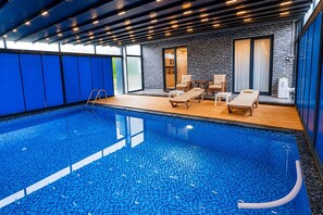Een binnenzwembad met helderblauw water, een houten terras, ligbedden en een bakstenen muur.