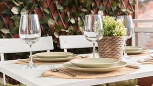 Una mesa de comedor dispuesta con platos, cubiertos y copas de vino, con una cesta tejida que contiene follaje en el centro.