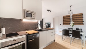 Una cocina moderna con horno empotrado, microondas y cafetera. Hay un comedor con una mesa dispuesta para cuatro y una silla.
