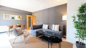 Ein modernes Wohnzimmer mit einem grauen Sofa, einem Glastisch, einem schwarzen Beistelltisch und einer Zimmerpflanze.