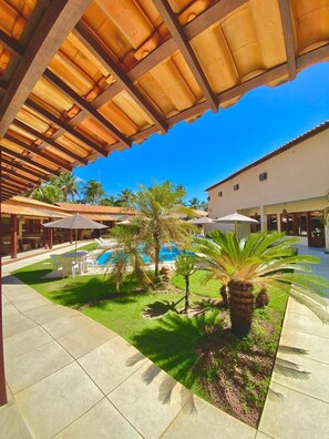 A covered outdoor area with a pool, palm trees, and a building with a tiled roof.