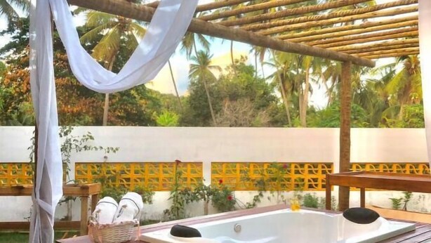 Un jacuzzi de madera bajo una pérgola con cortinas blancas, rodeado por un jardín con palmeras y una valla amarilla.