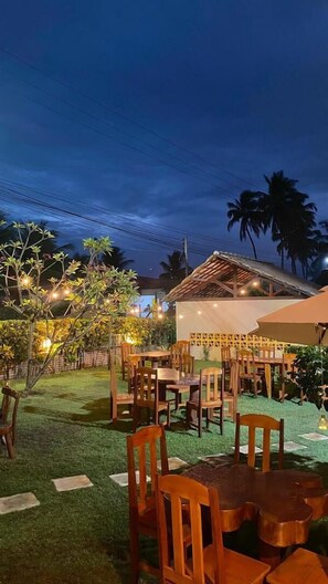 An outdoor dining area with wooden tables and chairs, surrounded by greenery and palm trees.