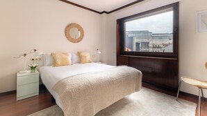 Un dormitorio con una cama grande, una mesita de noche con lámpara, una ventana con vistas a edificios y un espejo redondo en la pared.