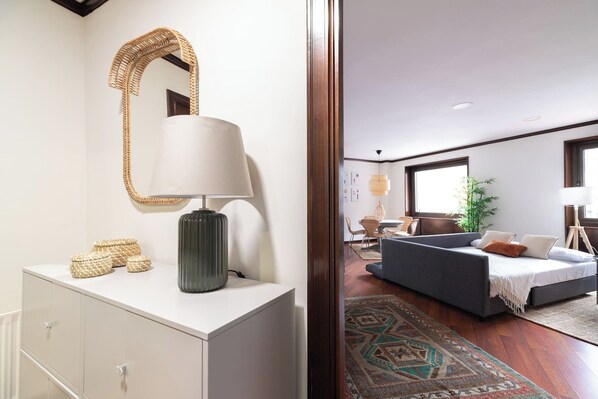 Une pièce avec un meuble blanc, un miroir à motifs tissés, une lampe et un canapé gris agrémenté d’un tapis à motifs.