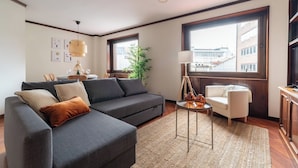 Una sala moderna con un sofá seccional gris, un sillón blanco pequeño, una mesita de centro redonda y un ventanal con vistas a edificios.