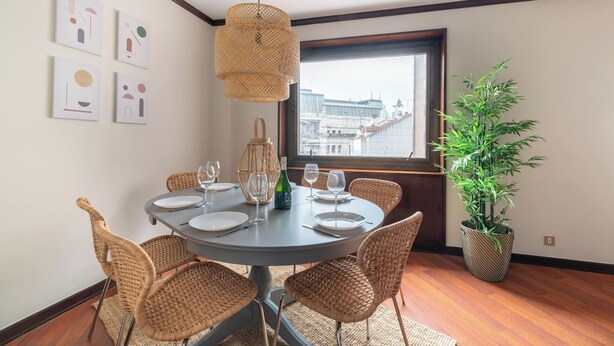 Comedor con una mesa redonda dispuesta para cuatro personas, rodeada de sillas de mimbre. Se observa un ventanal con vistas a edificios, una planta en maceta y una obra de arte en la pared.