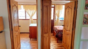 A room with wooden flooring, a bed with a patterned bedspread, a wooden wardrobe, and a window with curtains.