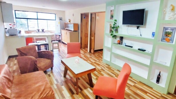 A living room with a wooden floor, a TV mounted on a green wall, and a kitchen area in the background.