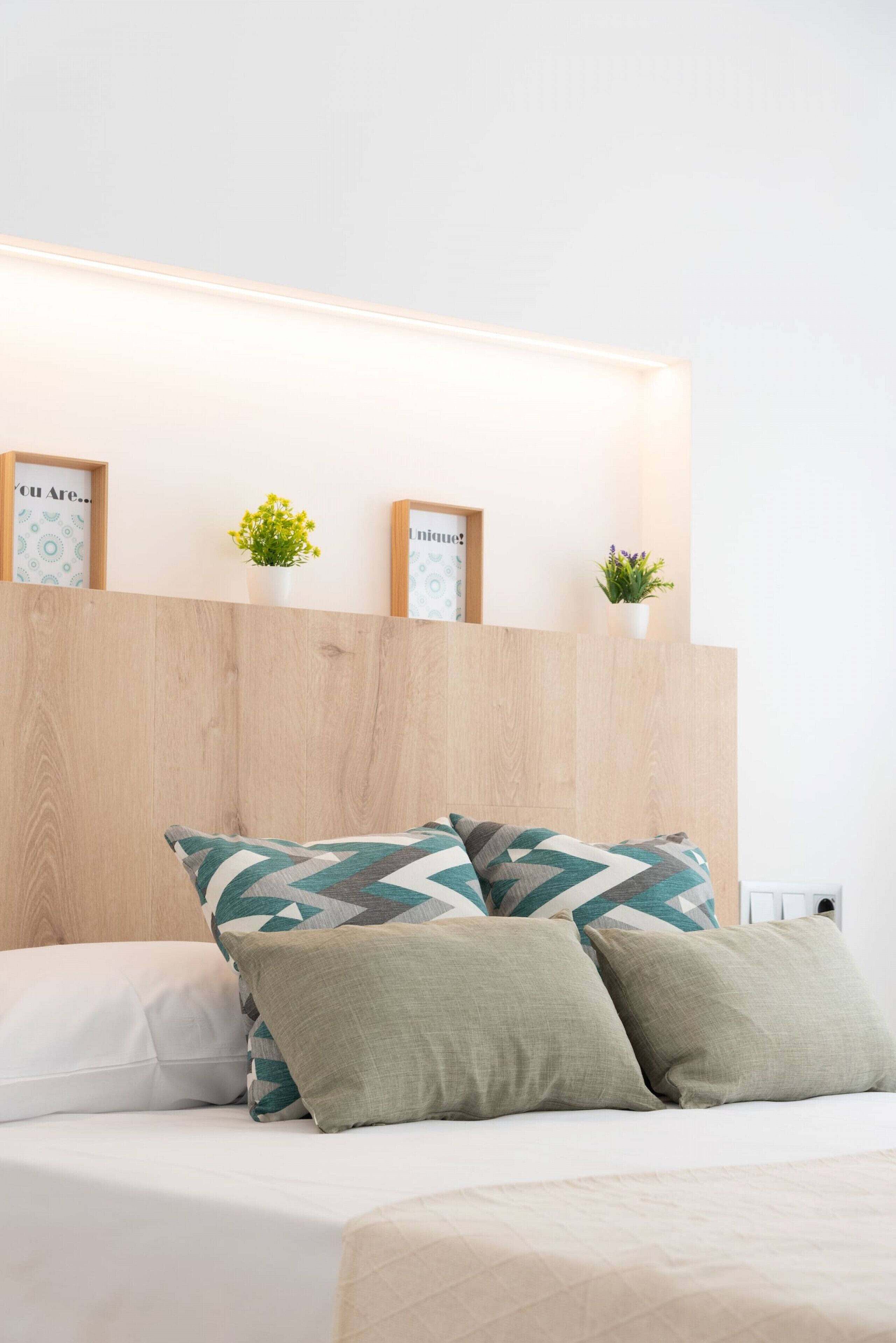 A neatly made bed with a geometric patterned pillow, a plain pillow, and a beige pillow. There are two small potted plants on a wooden shelf above the bed.