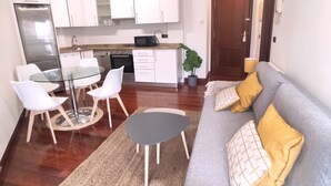 A modern living room with a grey sofa, a glass dining table, and white chairs. A kitchen with white cabinets and a microwave is visible in the background.