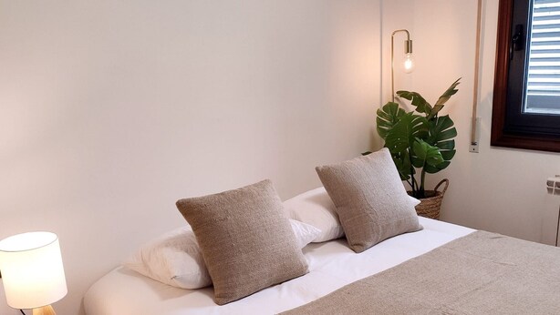 A neatly made bed with a beige blanket and pillows, a bedside lamp, and a potted plant.
