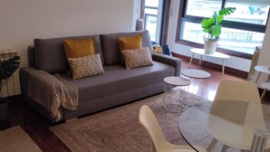 A modern living room with a grey sofa, two yellow cushions, a glass coffee table, and a potted plant by the window.
