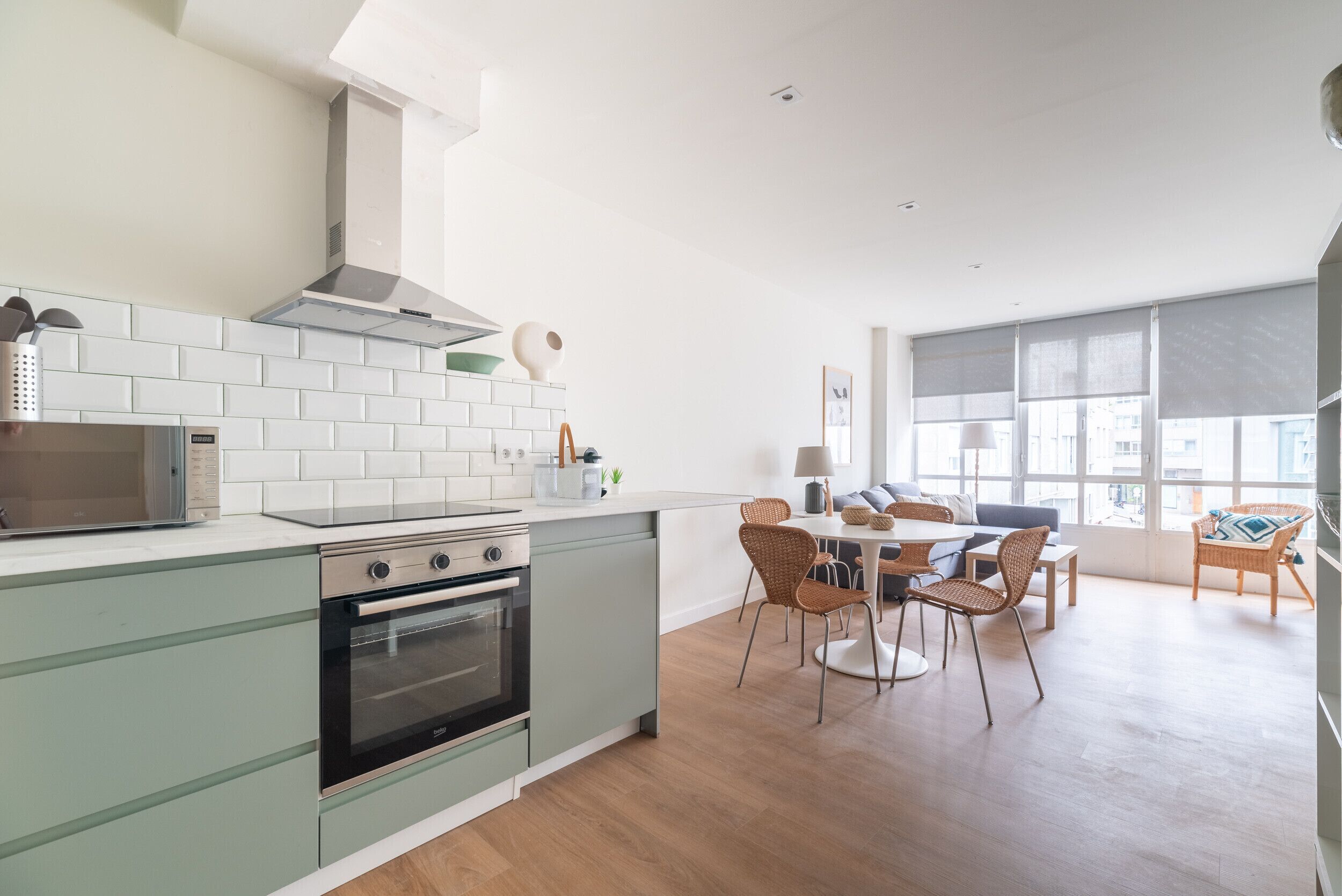 A modern kitchen with white subway tiles, a stainless steel range hood, and a built-in oven. There is a dining area with a table and chairs, and large windows providing natural light.
