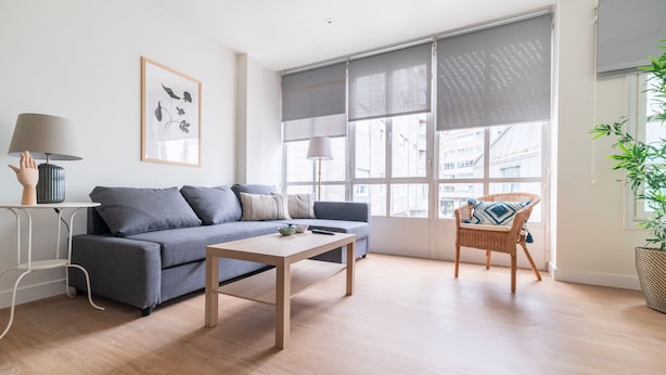 Un salón moderno con un sofá gris, una mesa de centro de madera, una silla de mimbre y un amplio ventanal con persianas.