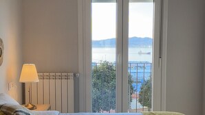 Quarto com cama, mesa de cabeceira com abajur e vista para o mar através de uma porta de correr.