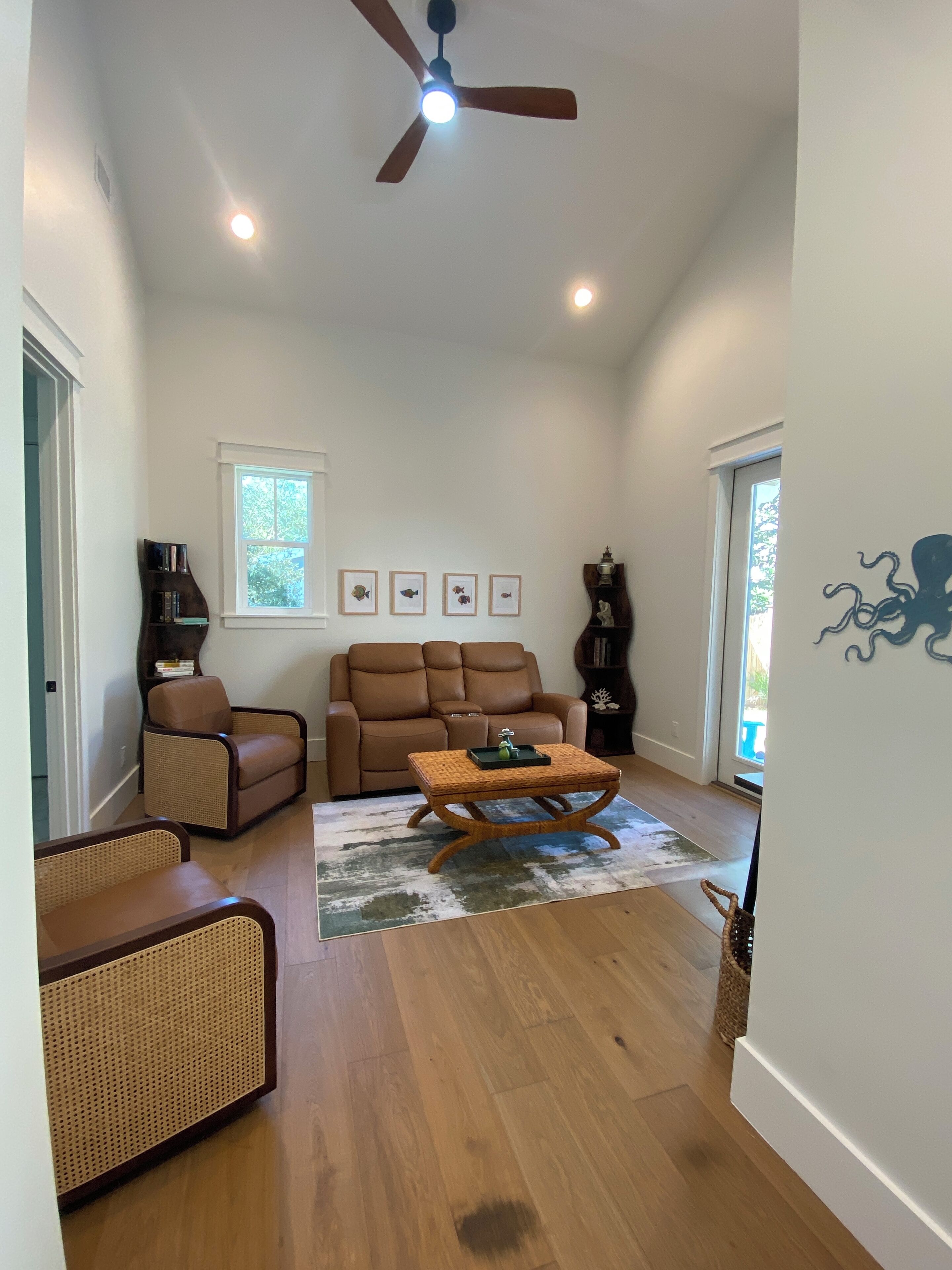 A spacious living room with a brown leather sofa, a wicker chair, a wooden coffee table, and a ceiling fan.