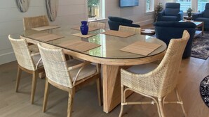 A dining area with a glass table surrounded by wicker chairs, a chandelier, and a television in the background. - Beach Bum Luxury 30A, Heated Pool, Golf Cart, Bikes, 3 Minute Walk to The Beach! (Santa Rosa Beach)