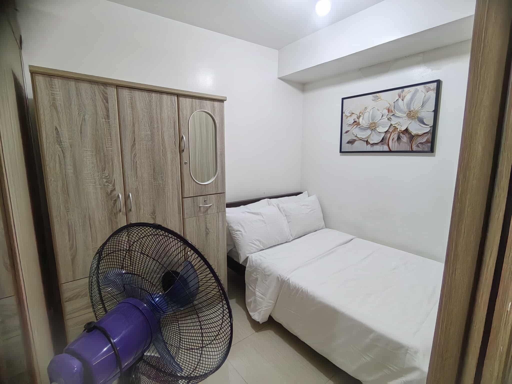 Une chambre à coucher avec un lit, une armoire, un ventilateur et un tableau floral accroché au mur.