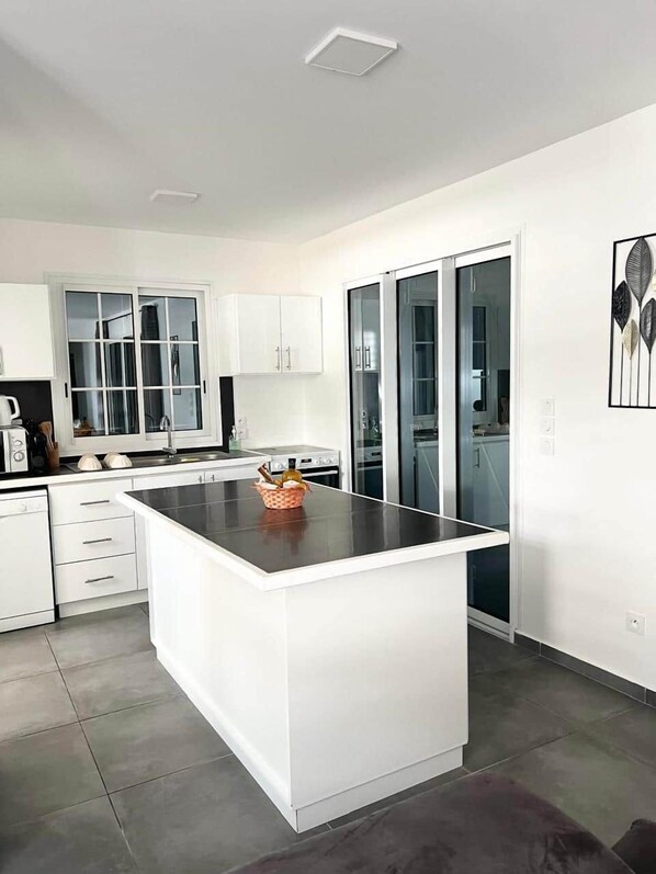 A modern kitchen with white cabinetry, a central island, and a glass door leading to a dining area. - Le Roc de Malendure (Bouillante)
