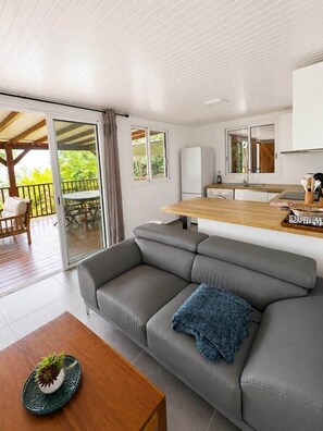 A modern living room with a grey sofa, a wooden coffee table with a decorative plate, and a kitchen area with a wooden countertop and white cabinets. - Le Roc de Malendure (Bouillante)