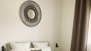 A modern bedroom with a bed, nightstands, a wall-mounted mirror, and a window with curtains. - Le Roc de Malendure (Bouillante)