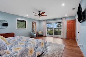 A bedroom with a bed, a ceiling fan, a TV, a window, and a door leading to a balcony.