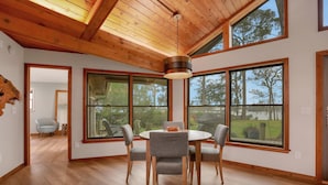 Zona pranzo con tavolo rotondo e quattro sedie, ampie finestre e soffitto in legno.