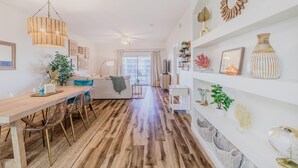 A modern living room with a wooden dining table, clear chairs, a sofa, and a ceiling fan.