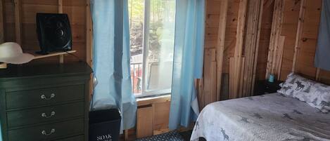 A wooden cabin room with a bed, a dresser, a fan, and a window with curtains.