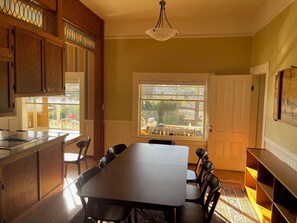 A dining area with a long table and chairs, wooden cabinets, a chandelier, and a large window with a view of the outdoors.