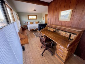 A room with a wooden desk, a bed with white bedding, and a window with blinds.