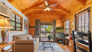 A cozy cabin interior with a wooden ceiling, a ceiling fan, a sofa, a coffee table, a TV, and a fireplace.