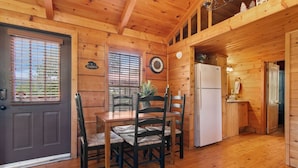 A wooden cabin interior with a dining table, chairs, a refrigerator, and a clock on the wall.