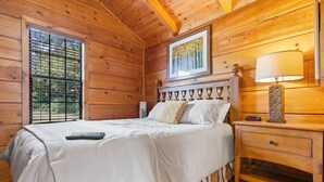 A wooden cabin room with a bed, a nightstand, a lamp, and a window with blinds.