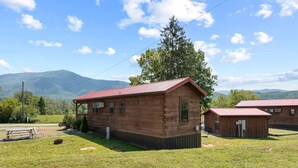 Un rustico chalet in legno con tetto rosso, un tavolo da picnic e un piccolo deposito in un prato erboso, con montagne sullo sfondo.