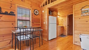 Interno di una baita in legno con un tavolo da pranzo, sedie, un frigorifero e una sveglia appesa al muro.