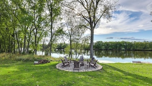 A circular outdoor seating area with Adirondack chairs and a fire pit, surrounded by a grassy lawn and trees, with a lake in the background. - Riverfront Home w/ Private Dock in Wisconsin Dells (Wisconsin Dells)