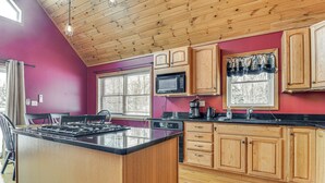 Una cocina con armarios de madera, encimera negra, microondas y ventana con persianas.