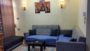 Ein Wohnzimmer mit grauem Sofa, einem kleinen Sessel, einem Couchtisch und gemusterten Teppichen. An den Wänden sind Leuchten angebracht und es gibt einen Deckenventilator.