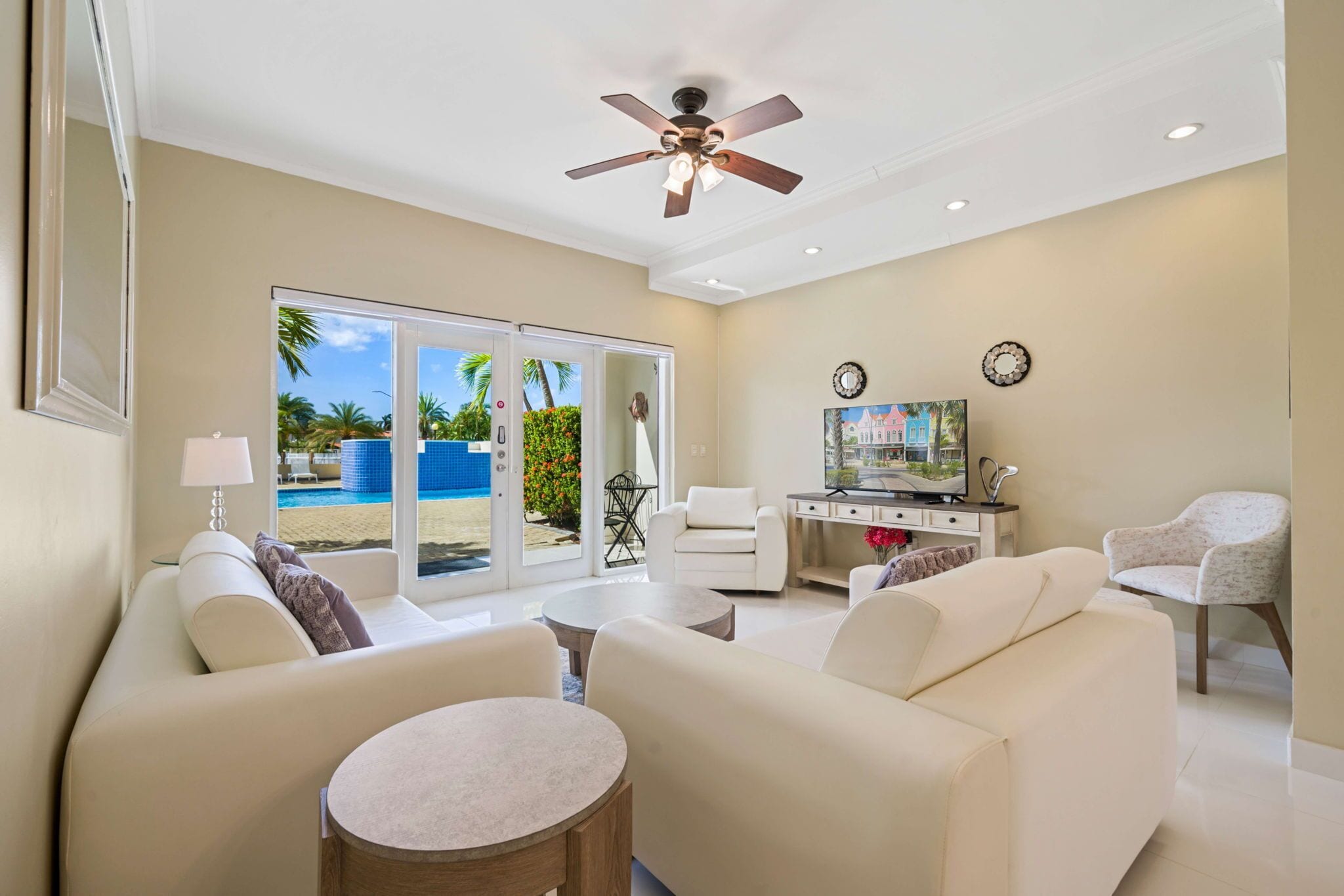 A spacious living room with a ceiling fan, a large window with a view of a pool, a white sofa, a coffee table, and a TV on a wooden stand.