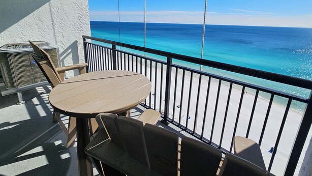 En balkon med et bord og stole, der giver udsigt over en strand med klart, blåt vand og en sandet kyst.