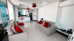 Una sala de estar moderna con dos sofás grises, cojines rojos, un televisor y una cocina al fondo.