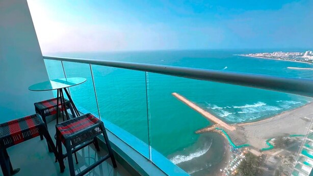 Un balcón con una mesa y sillas de vidrio, con vista a una zona costera de aguas azul turquesa y una playa de arena.