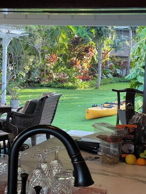 A view through a window of a room with a table, a chair, and a view of a garden with various plants and a yellow kayak. - Luxury Lanikai Oceanfront Home! (Kailua)