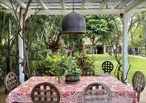 A covered outdoor dining area with a table set for four, surrounded by lush tropical plants and a pergola overhead. - Luxury Lanikai Oceanfront Home! (Kailua)