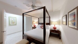 Una cama con dosel, un ventilador de techo, dos mesitas de noche con lámparas, una puerta y cuadros en las paredes.