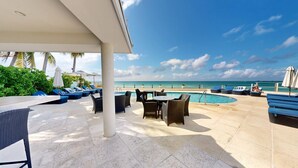Un espace aménagé au bord de la piscine, avec des chaises longues, des tables et des parasols, offrant une vue sur une plage sous un ciel dégagé.