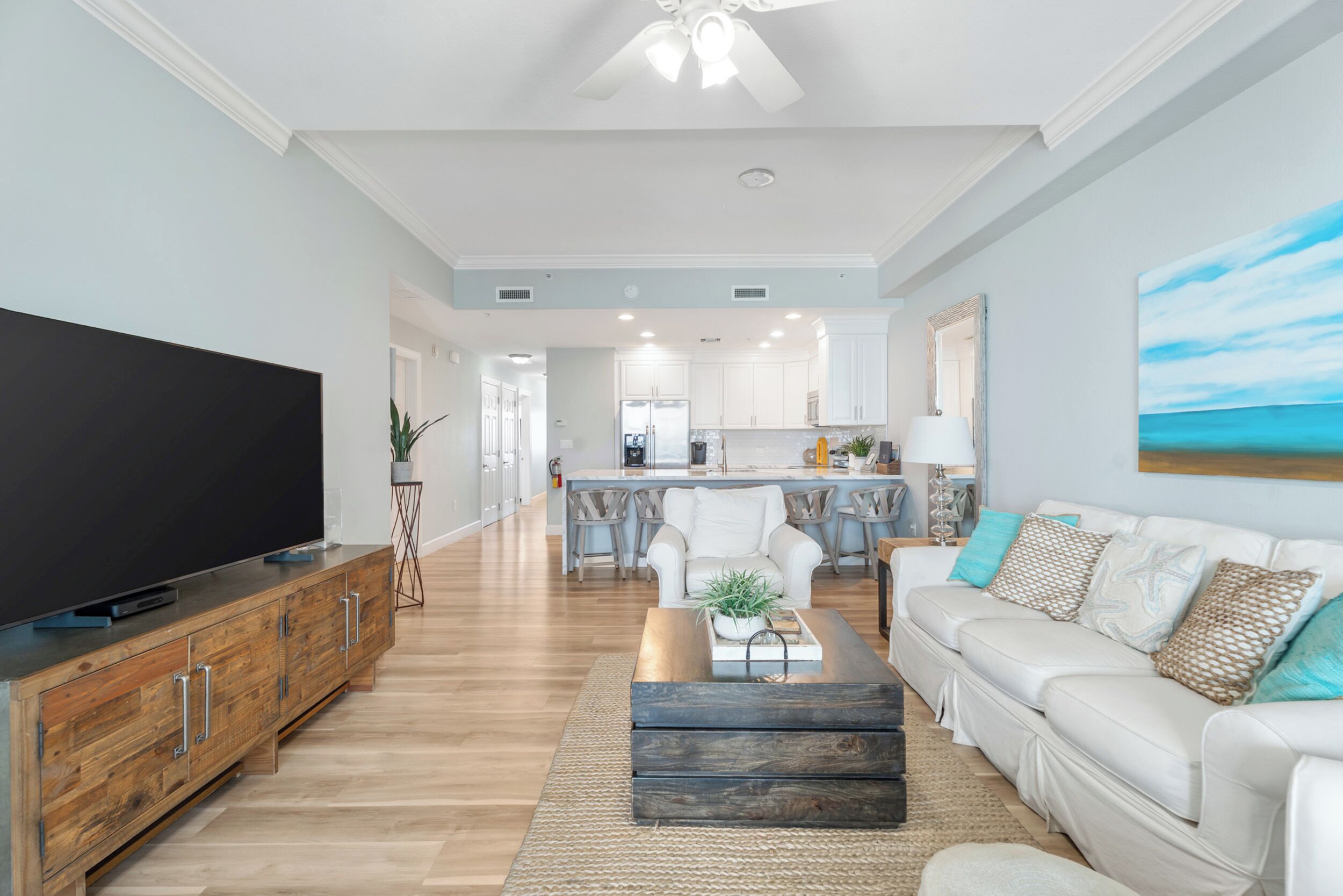 Una sala moderna con un televisor de pantalla plana, un mueble de madera, un sofá blanco y una cocina al fondo.